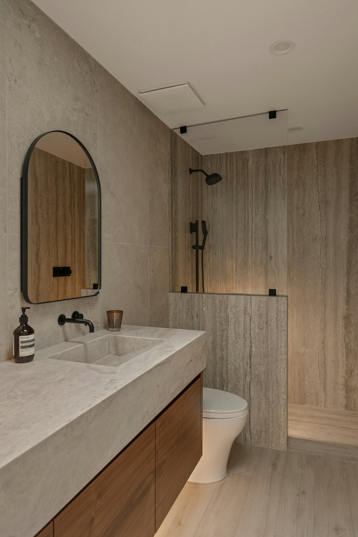 Baño con paredes de piedra clara, mueble de madera y espejo en arco
